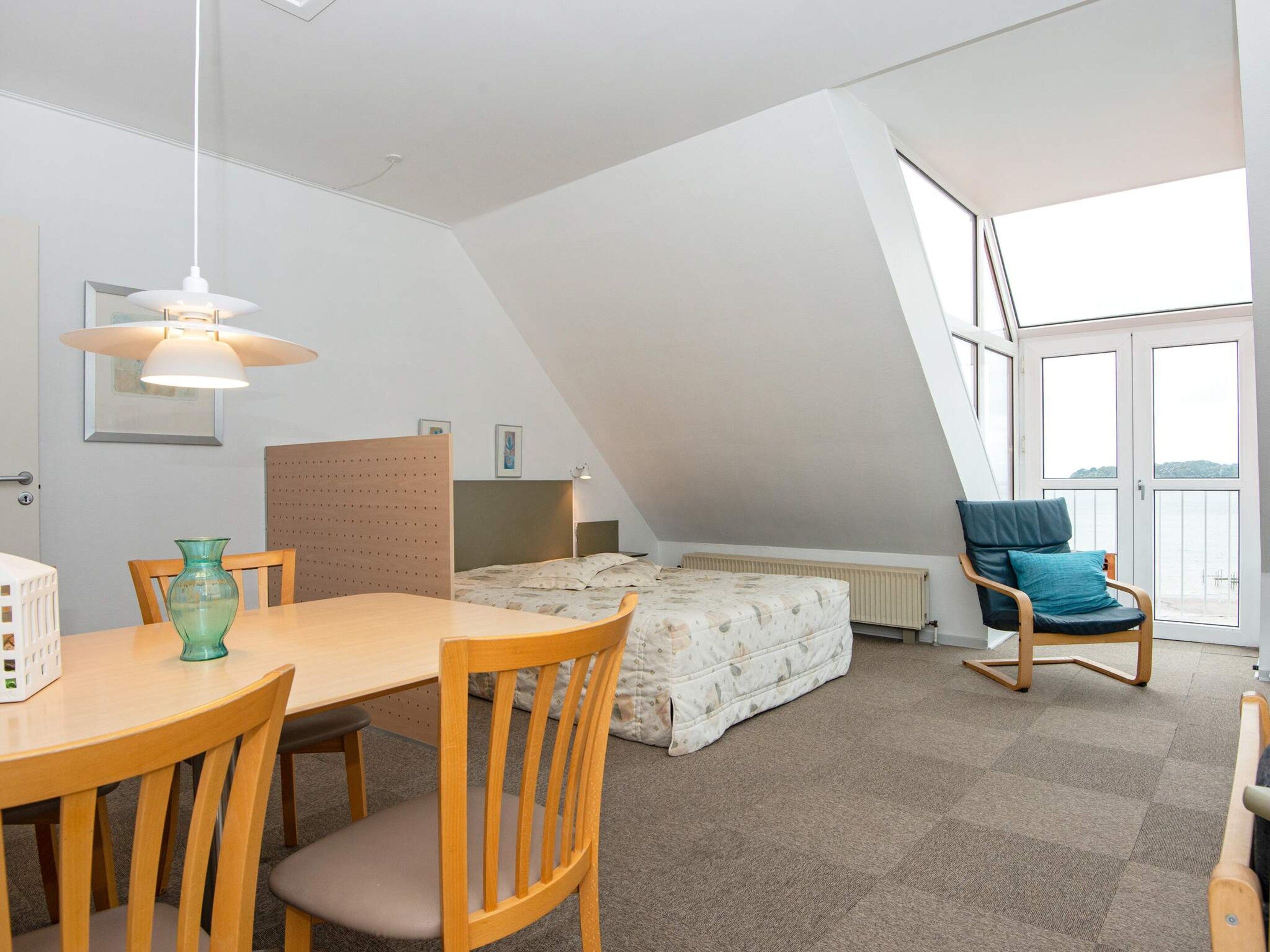 A room with a dining table, chairs, a bed, a window, and a chair with a cushion.