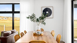 Un comedor con una mesa de madera, sillas de mimbre, un jarrón con follaje y una obra de arte enmarcada en la pared.