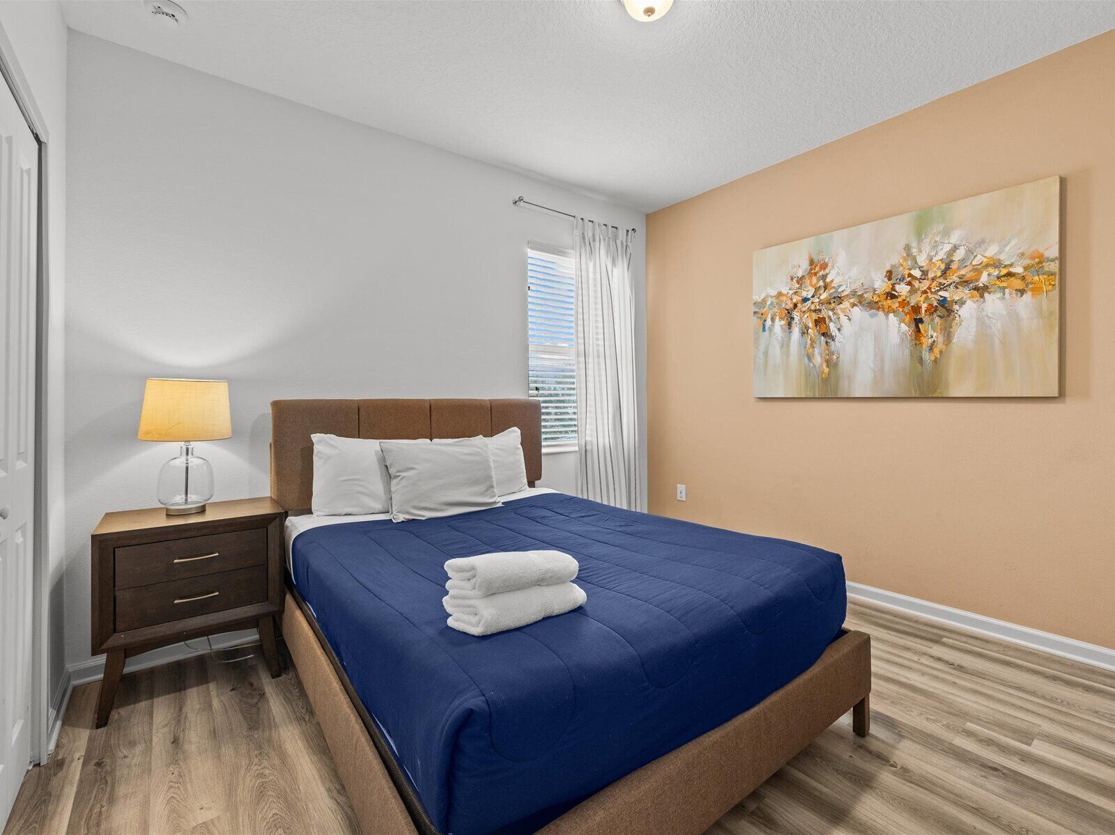 A neatly arranged bedroom with a bed, bedside table, lamp, and a painting on the wall.