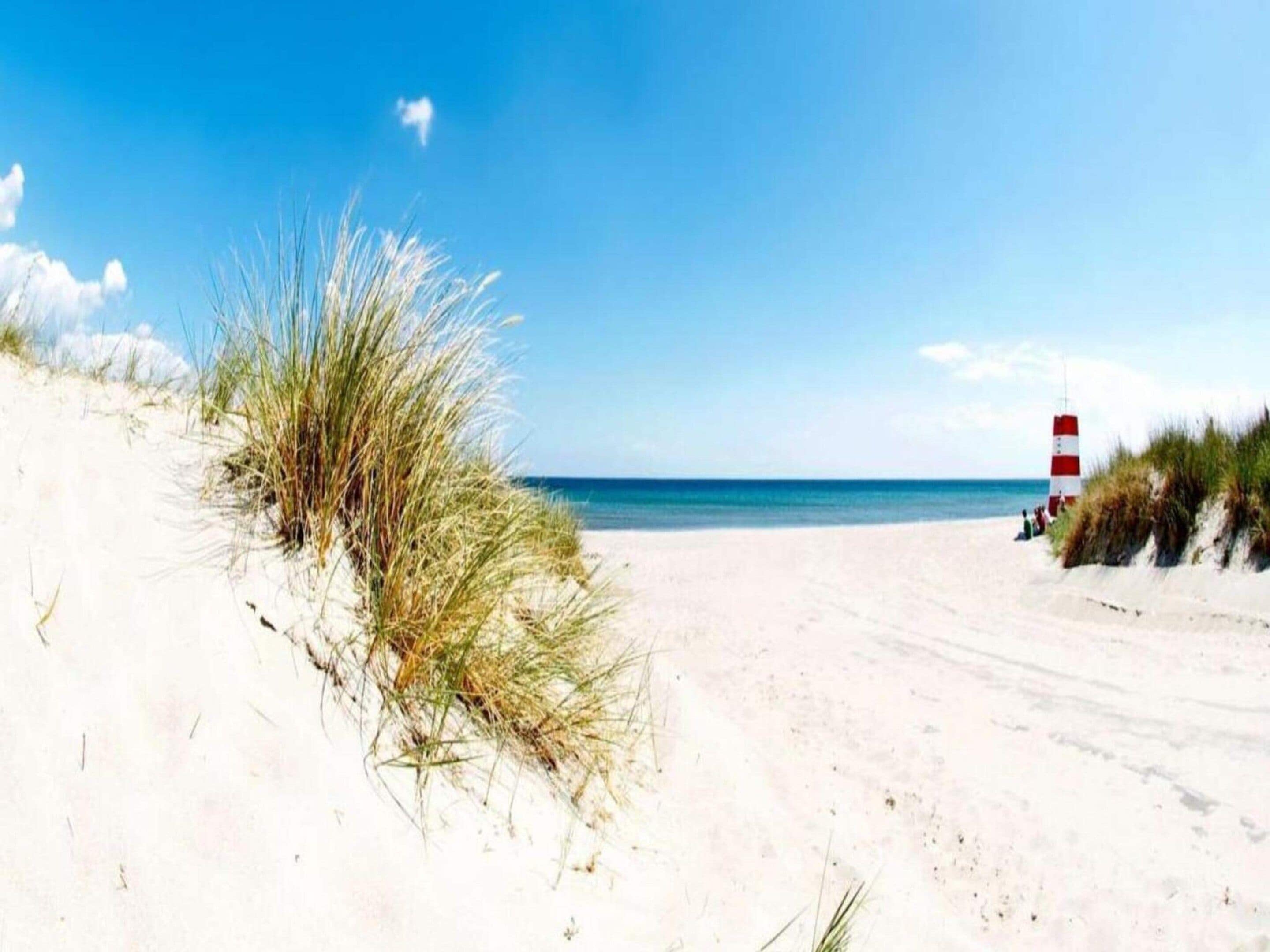 A beach with sand dunes, a clear sky, and a red and white lighthouse in the distance.