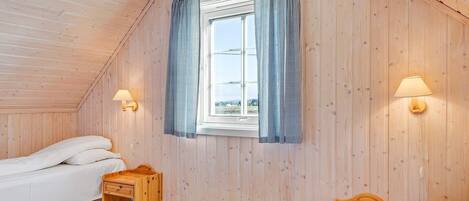 Een knus vertrek met houten wanden, een bed, een klein houten tafeltje, een nachtkastje en een raam met geruit gordijnen.