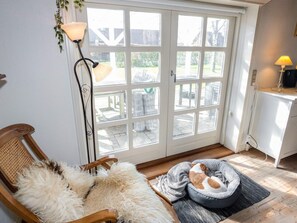 A room with a large glass door leading outside, a wooden chair with a furry throw, a pet bed, and a lamp.