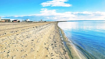 Una amplia playa de arena con huellas que conducen al agua, un cielo despejado y edificios a lo lejos.