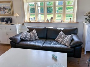 A modern living room with a black leather sofa, a white coffee table, and a window with multiple panes overlooking a garden. - 5 Person Holiday Home in Oster Assels (Øster Assels)