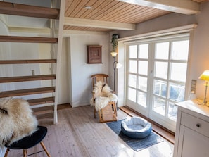 A room with a wooden staircase, a fluffy dog on a chair, a dog bed, a window with a view, and a lamp on a table.