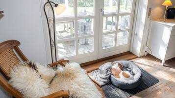 Una habitación con un gran ventanal que da al exterior, una silla de madera con una manta mullida, una cama para mascotas color gris y una mesita con una lámpara.