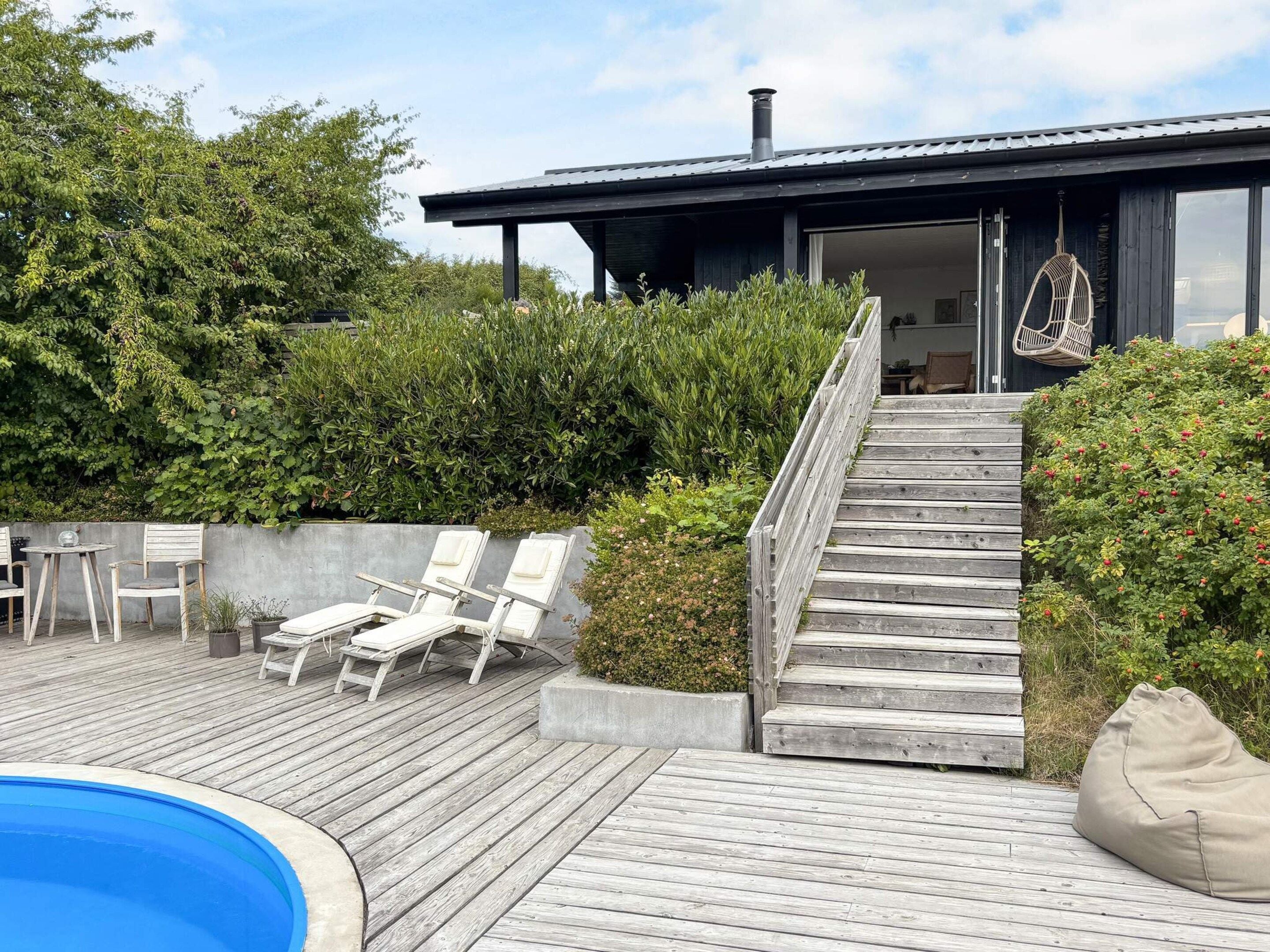 Una terraza de madera con sillones de descanso, una piscina y una escalera que conduce a un edificio con chimenea.