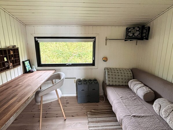A compact room with a wooden desk, a white chair, a sofa, a window with a view of greenery, and a small shelf with decorative items. - 4 Star Holiday Home in Ebeltoft (Ebeltoft)