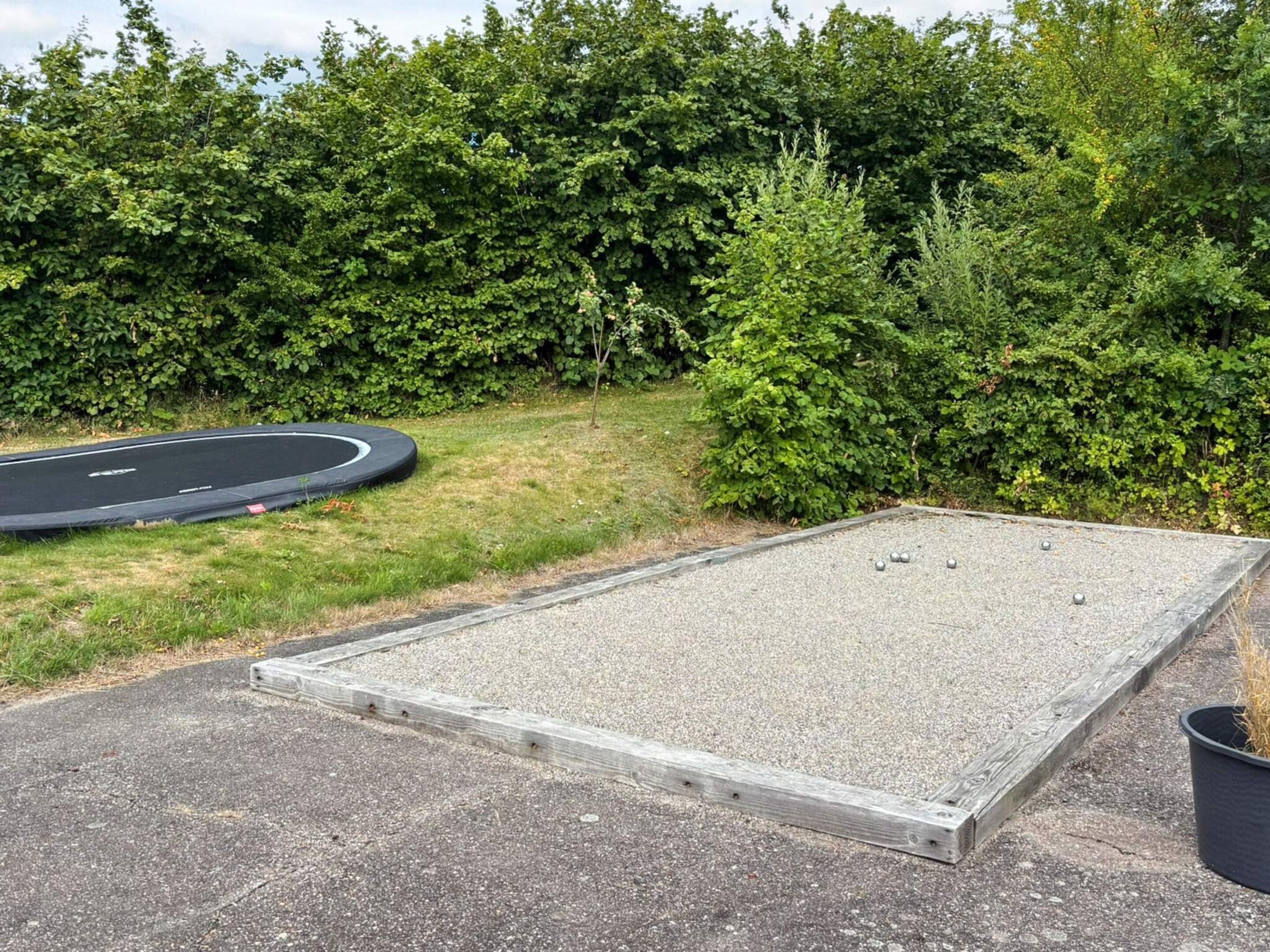 A rectangular outdoor area with a gravel surface, surrounded by a hedge and a circular trampoline.