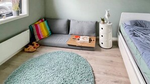 A cozy room with a grey sofa, a colorful striped pillow, a small white nightstand, a blue rug, and a bed with a grey blanket.