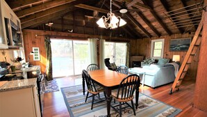 L’intérieur d’un chalet rustique comprend une table à manger en bois, des chaises, un coin cuisine, un canapé et une grande fenêtre.