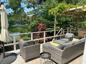 A deck with wicker furniture, a table, and an umbrella. - Waterfront Cottage Nestled In Wooded Privacy (2297) (Eastham)