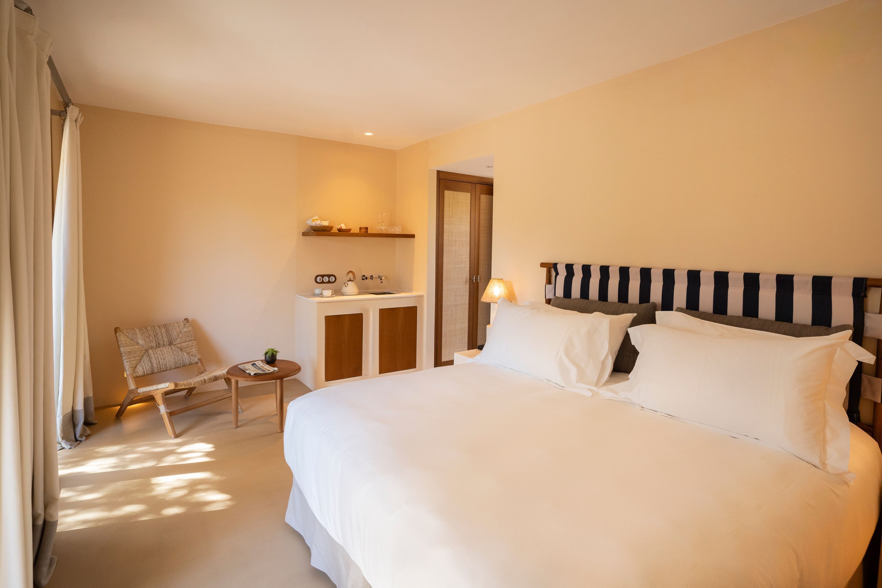 A hotel room with a large bed, a small desk with a chair, a nightstand, and a window with curtains.