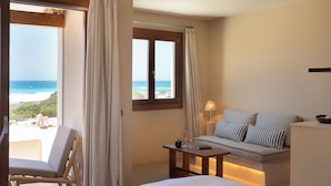 A room with a bed, a sofa with striped pillows, a small table with a lamp, and a view of the ocean through a window. - Dunas de Formentera Hotel (Formentera)