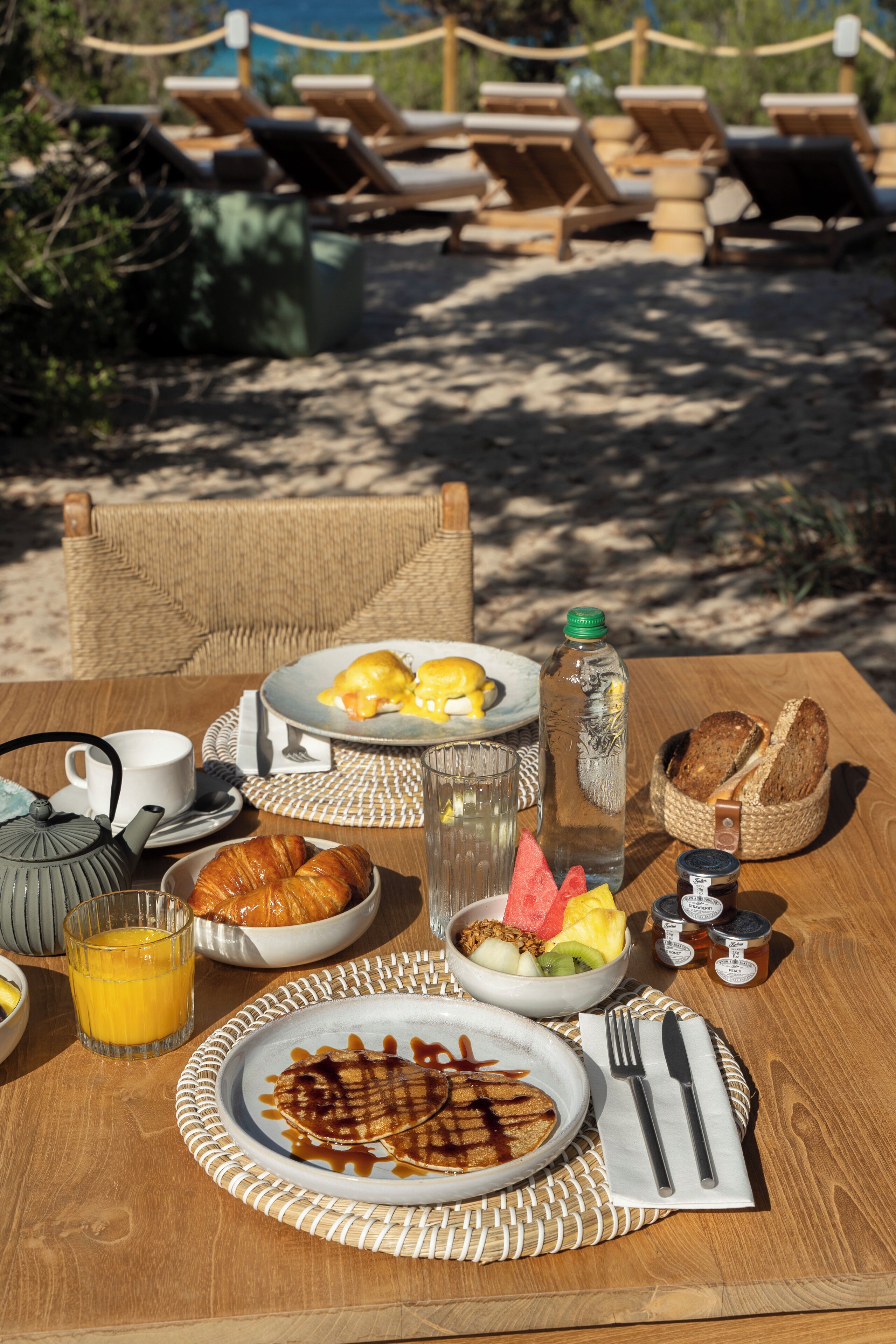 A breakfast table with croissants, eggs, and juice, set on a wooden table outdoors with lounge chairs in the background.