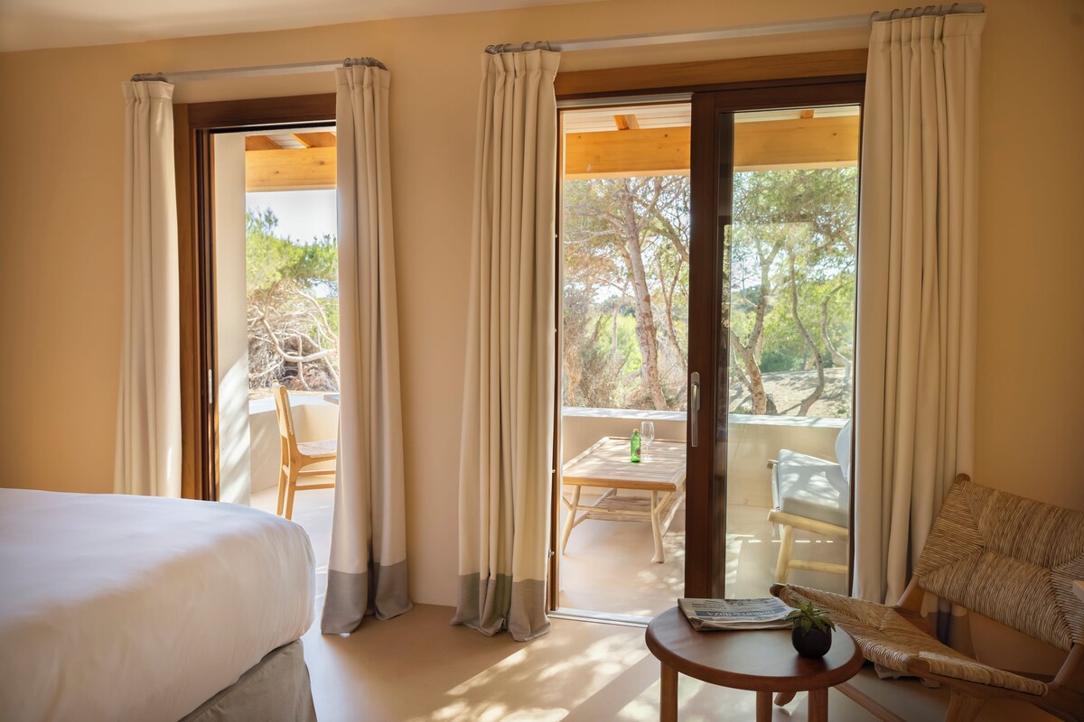 Une chambre d’hôtel avec un lit, une petite table sur laquelle se trouvent une bouteille et un verre, une chaise, et un balcon avec une table et des chaises.
