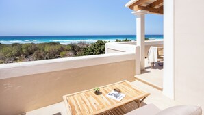 A balcony with a wooden table, a small plant, and a magazine. The balcony overlooks a beach with the ocean and greenery in the background. - Dunas de Formentera Hotel (Formentera)