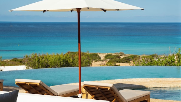 A poolside area with lounge chairs and a large umbrella, overlooking a clear blue sea and a distant coastline. - Dunas de Formentera Hotel (Formentera)
