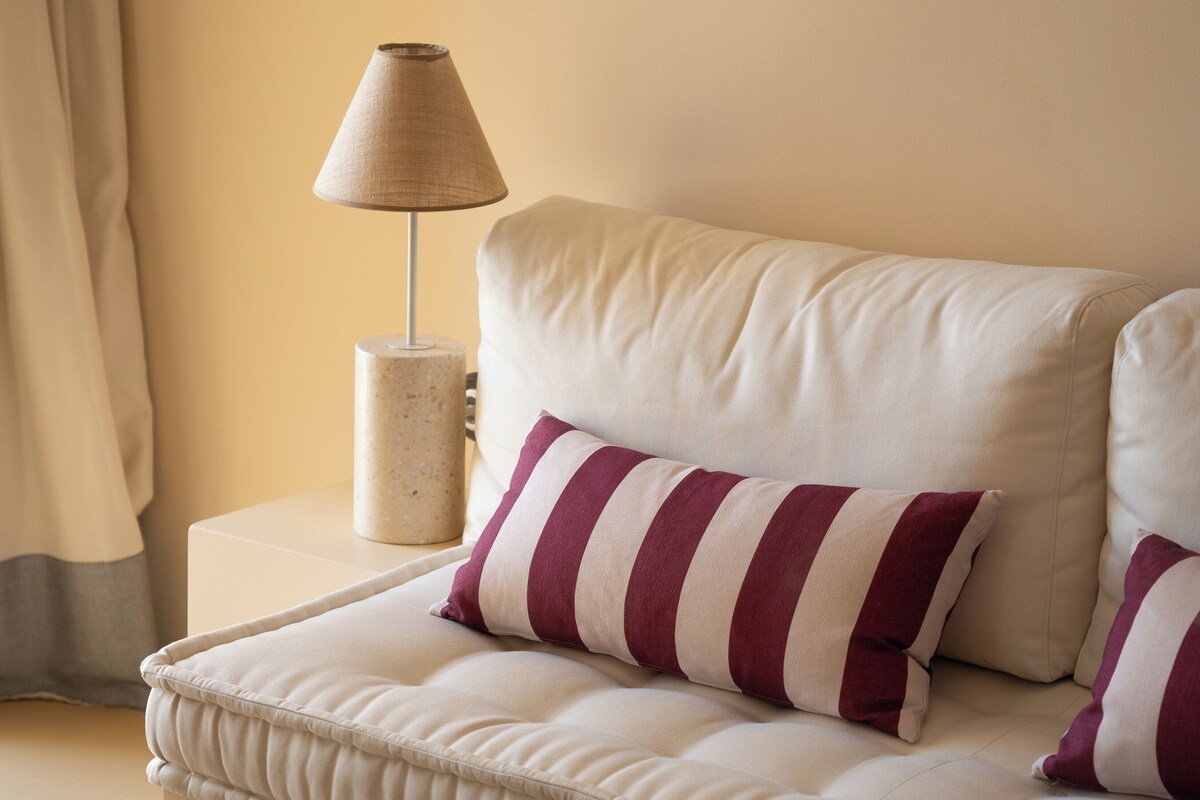 Habitación de hotel ordenada con sofá, almohadas a rayas, una lámpara y una mesita con papeles.