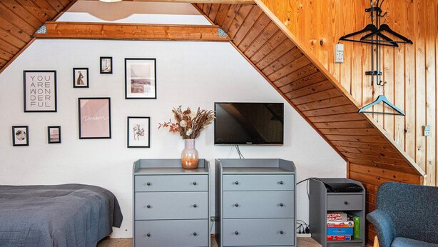 A bedroom with a sloped ceiling, a bed, two dressers, a TV, and wall art.