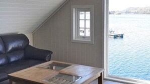 Une pièce avec un canapé en cuir foncé, une table basse en bois dotée de panneaux de verre, une fenêtre donnant sur un plan d’eau et des bateaux, et un plafond en pente.