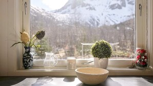 Een raam met uitzicht op besneeuwde bergen, een vaas met gedroogde bloemen, een kan, een mok, een klein potplantje en een schaal op een vensterbank.