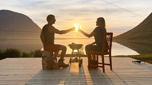 Twee personen zitten op een houten terras en kijken naar een zonsondergang boven het water, met bergen op de achtergrond.