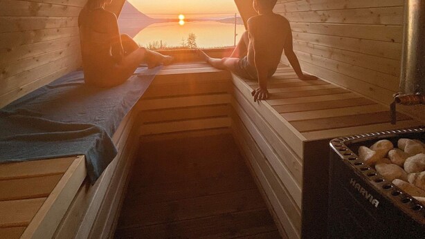 Een sauna met houten wanden en banken, uitzicht op de ondergaande zon en een sauna-badkuip, met zichtbaar het merk.