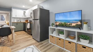 Une cuisine compacte avec des meubles blancs, un réfrigérateur en acier inoxydable, un coin repas avec une table et des chaises, et un téléviseur posé sur une étagère blanche, diffusant une image de plage.