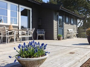 Une terrasse en bois avec des meubles d’extérieur, un pot de fleurs contenant des fleurs bleues et une maison dotée de grandes fenêtres et d’une porte.