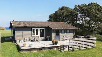 A small, single-story house with a tiled roof, a porch with outdoor furniture, and a view of the sea.