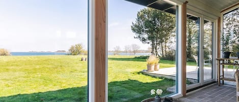 A covered patio with large windows overlooking a grassy field and a body of water.