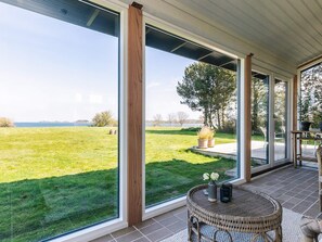 A covered patio with large windows overlooking a grassy field and a body of water.