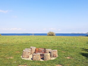 Ein grasbewachsenes Feld mit einer kreisförmigen Anordnung aus Holzstämmen und einem Metallzaun in der Mitte, mit Blick auf ein Gewässer.