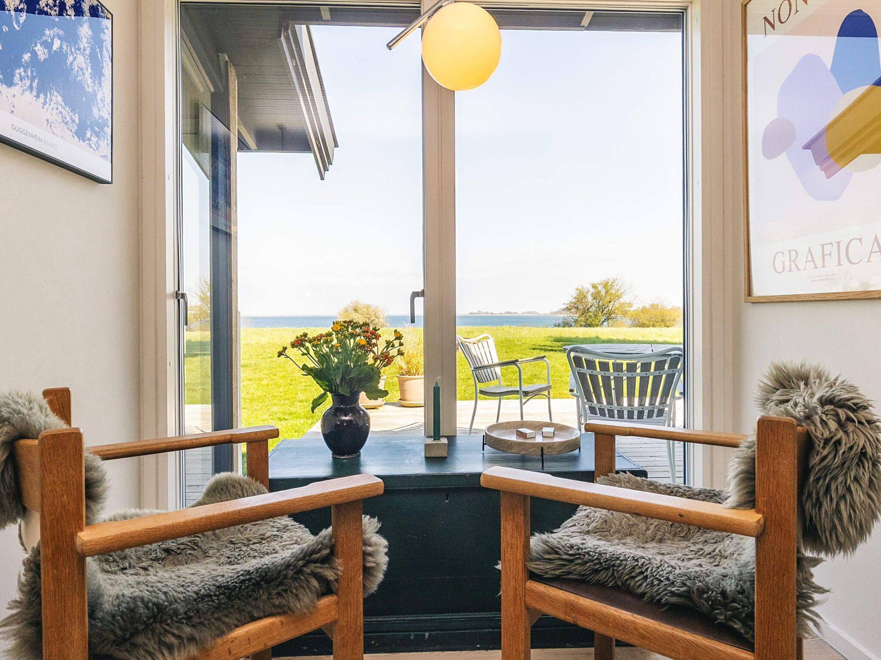 A room with two wooden chairs with fur throw cushions, a small table with a vase of flowers, and a view of a grassy field through a glass door.