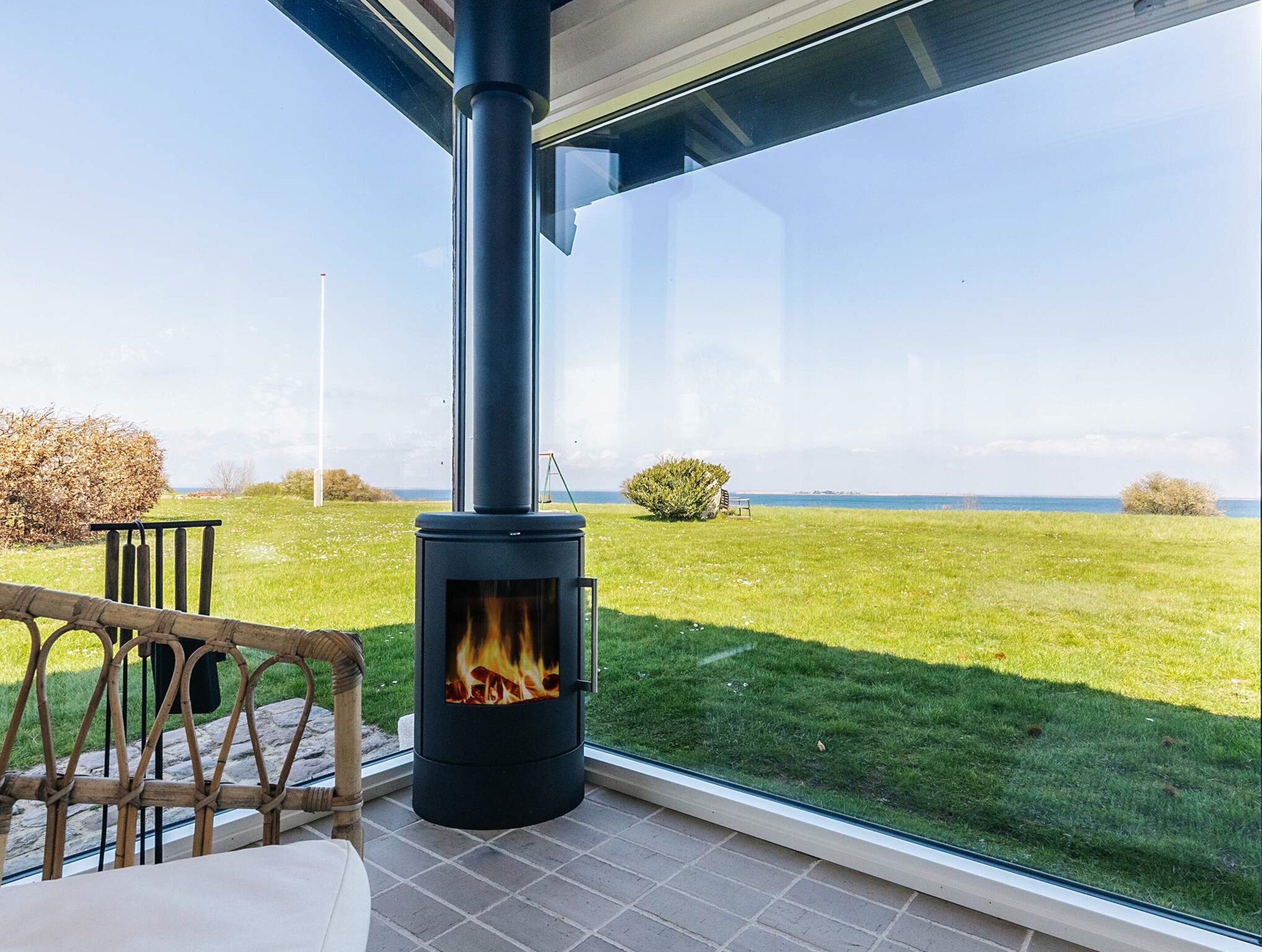 A modern outdoor patio with a wood-burning stove, a glass-enclosed seating area, and a view of a grassy field and water body.