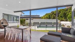 Un patio exterior moderno con terraza de madera, una mesa de comedor con sillas, un sillón colgante y una piscina visible a través de una valla.