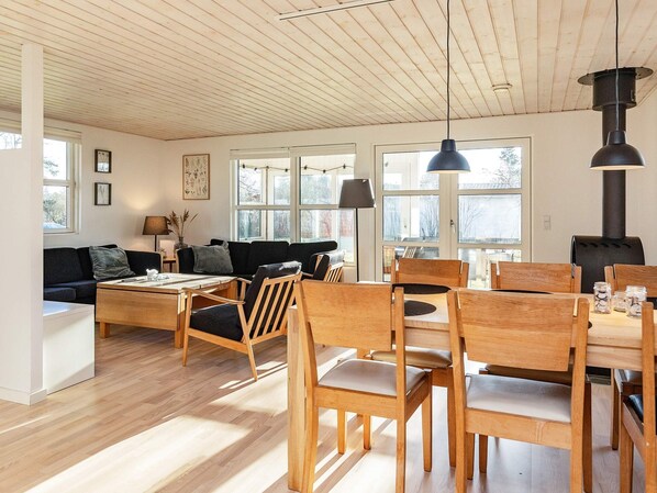A spacious living area with a wooden ceiling, a sofa set, a dining table with chairs, and a large window providing natural light.