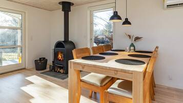 A modern interior with a wood-burning stove, a dining table with black placemats, and a view of the outdoors through large windows.