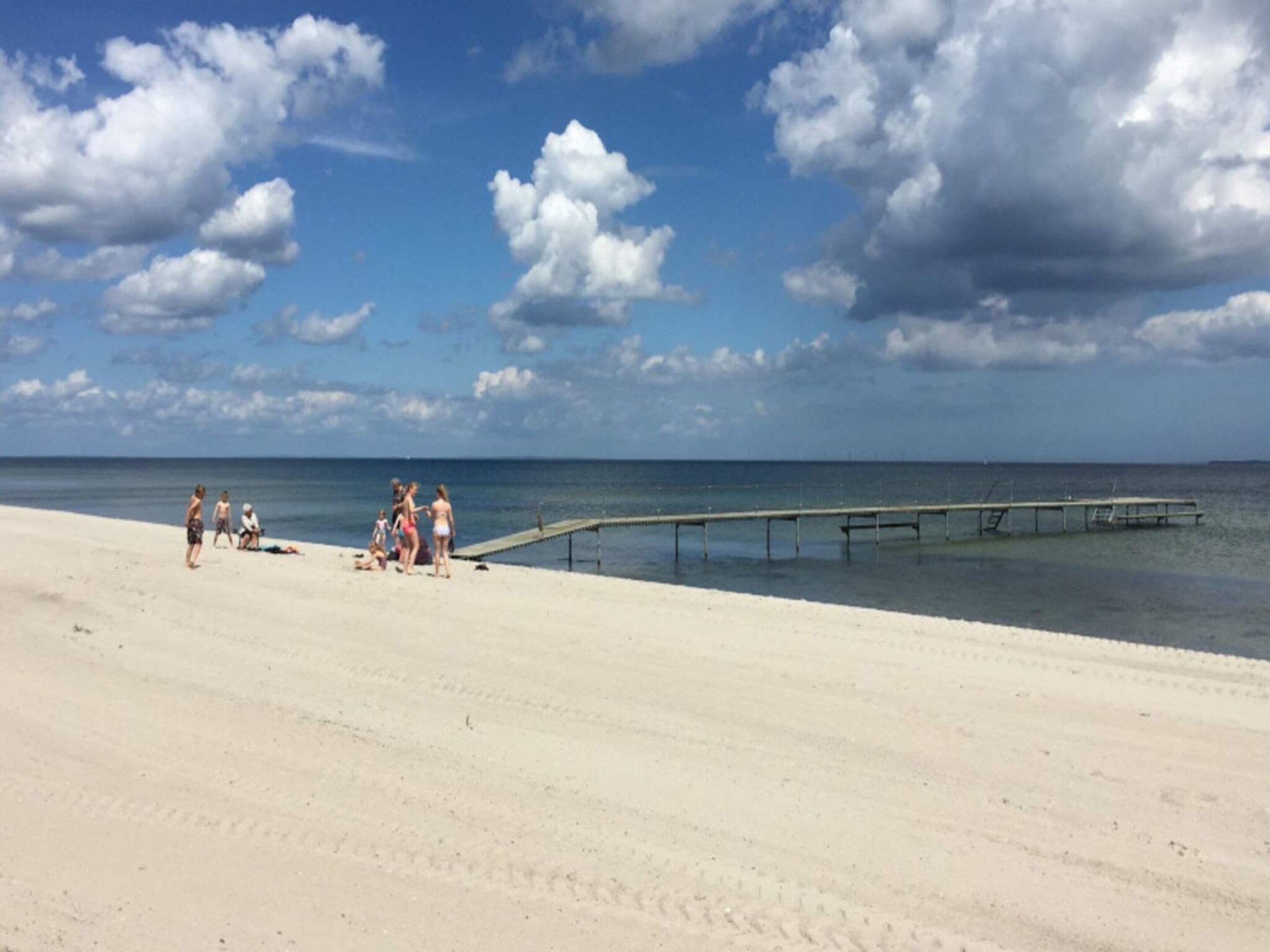 Una playa con un muelle de madera que se adentra en el agua, arena dorada y un cielo despejado con nubes.