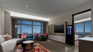 Habitación de hotel moderna con un ventanal grande que ofrece vistas a la ciudad, un televisor de pantalla plana montado en la pared y una zona de estar con una mesa de centro y sillas.