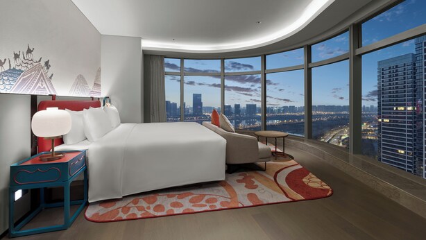 Habitación de hotel moderna con una cama grande, un área de descanso y vistas a la ciudad a través de amplios ventanales.