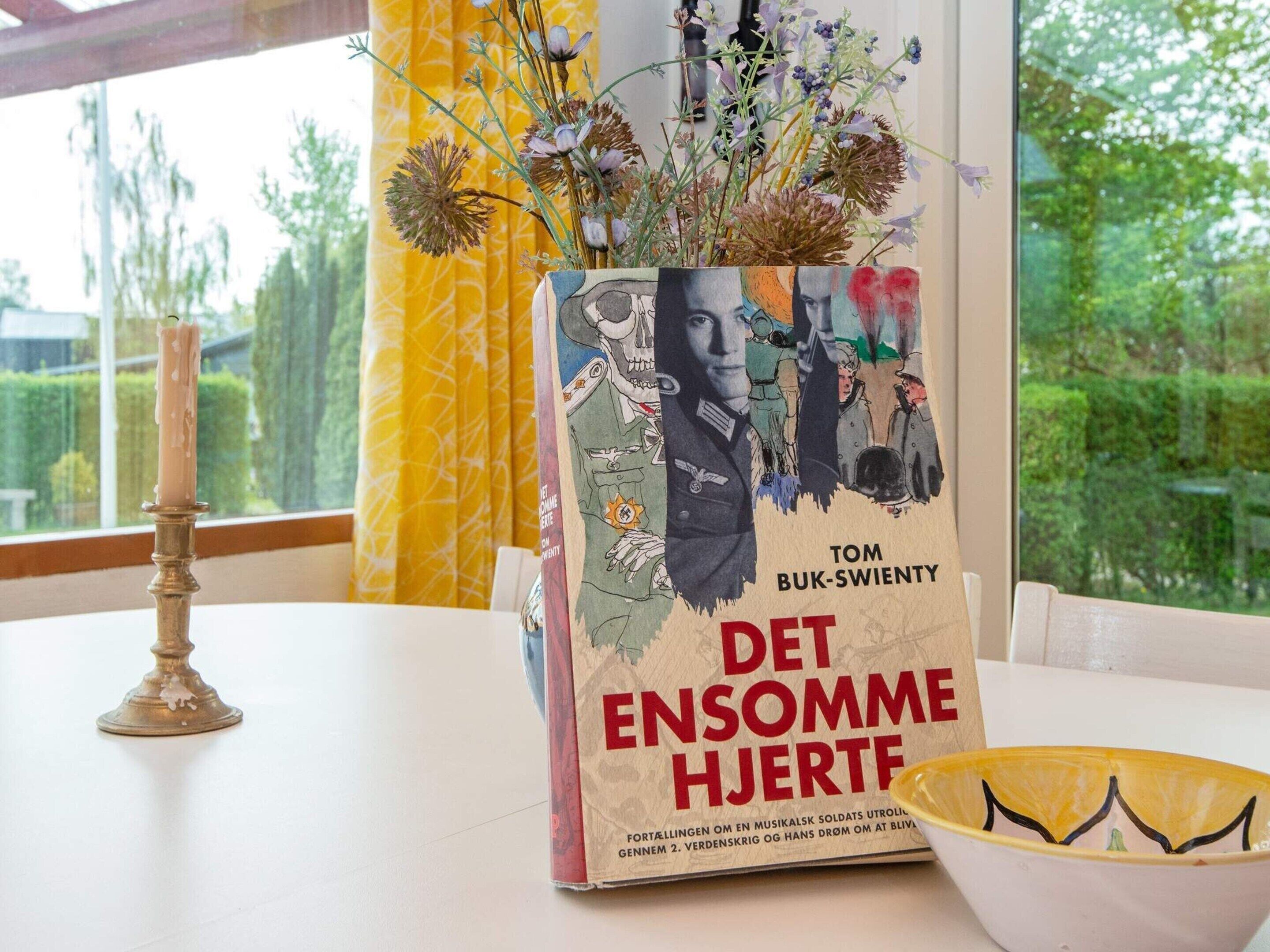 Un libro titulado “Det ensomme hjertes” de Tom Buk-Swenty, sobre una mesa con una vela y un cuenco decorativo.