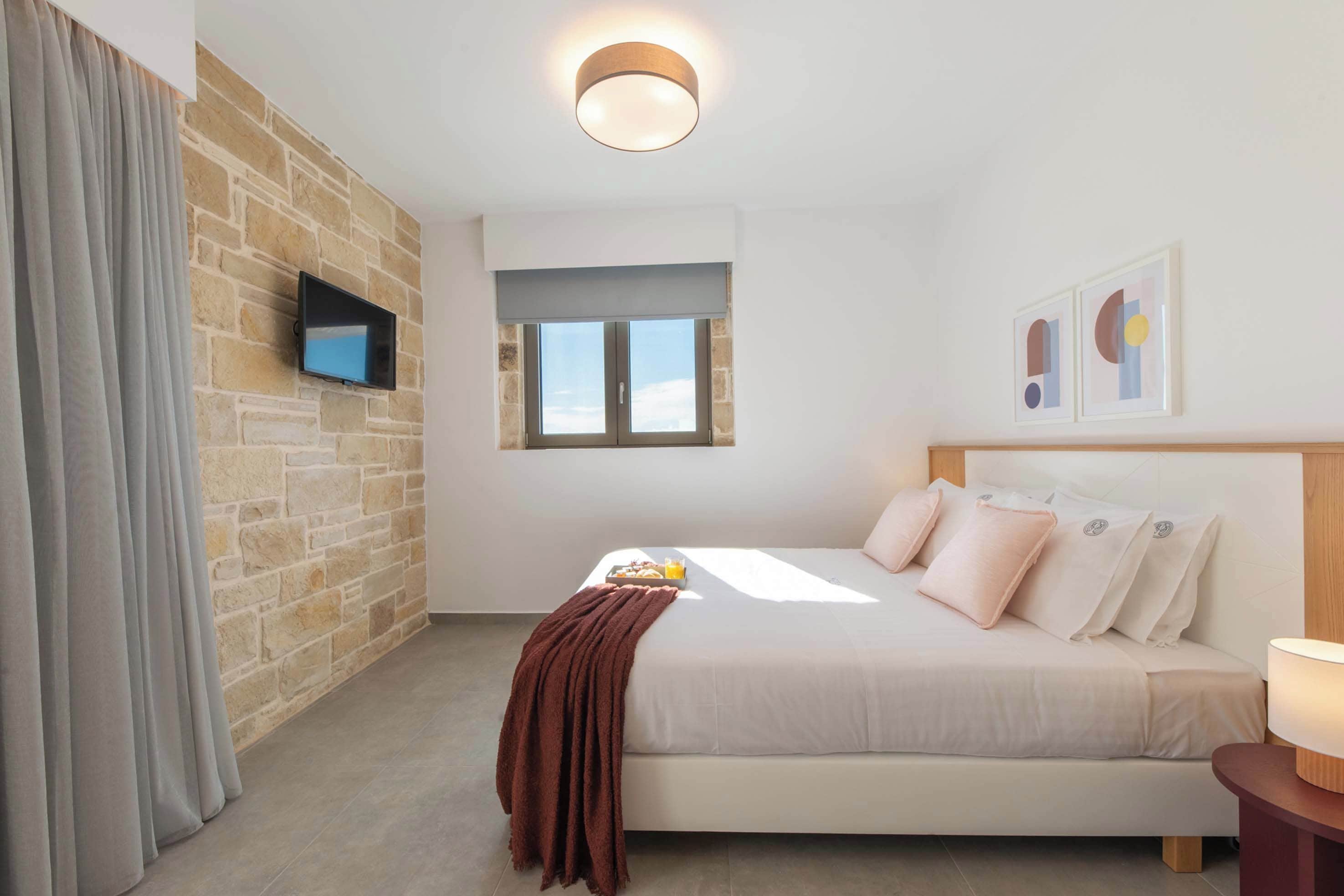 A modern bedroom with a stone wall, a flat-screen TV, a bed with pillows, a window, and a bedside table with a lamp.
