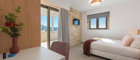 Habitación de hotel moderna con una cama blanca, un escritorio con un florero, una silla, un televisor montado en la pared y vistas de la ciudad a través de amplios ventanales.