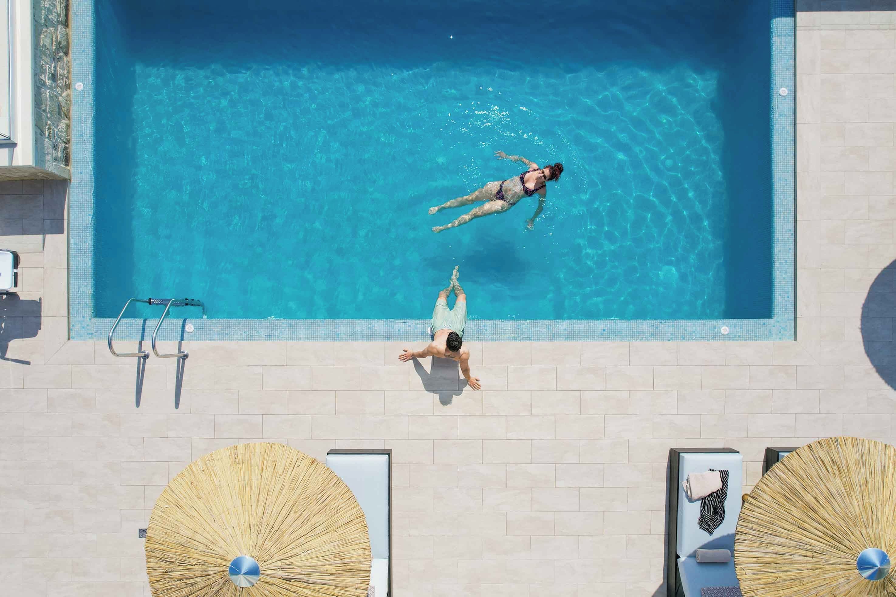 A swimming pool with two sun loungers and umbrellas, a ladder, and a tiled deck.