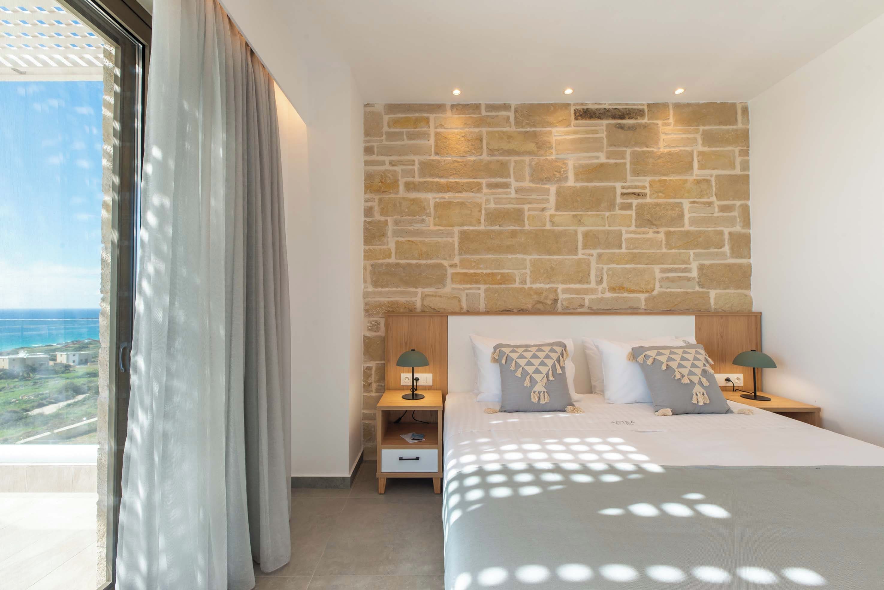 Une chambre avec un mur en pierre, un lit avec deux oreillers, une table de chevet et une vue sur l’océan.