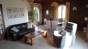A living room with a black leather sofa, a wooden coffee table, and a grey armchair. There are framed animal pictures on the wall, a dining table with chairs, and a large window with curtains.