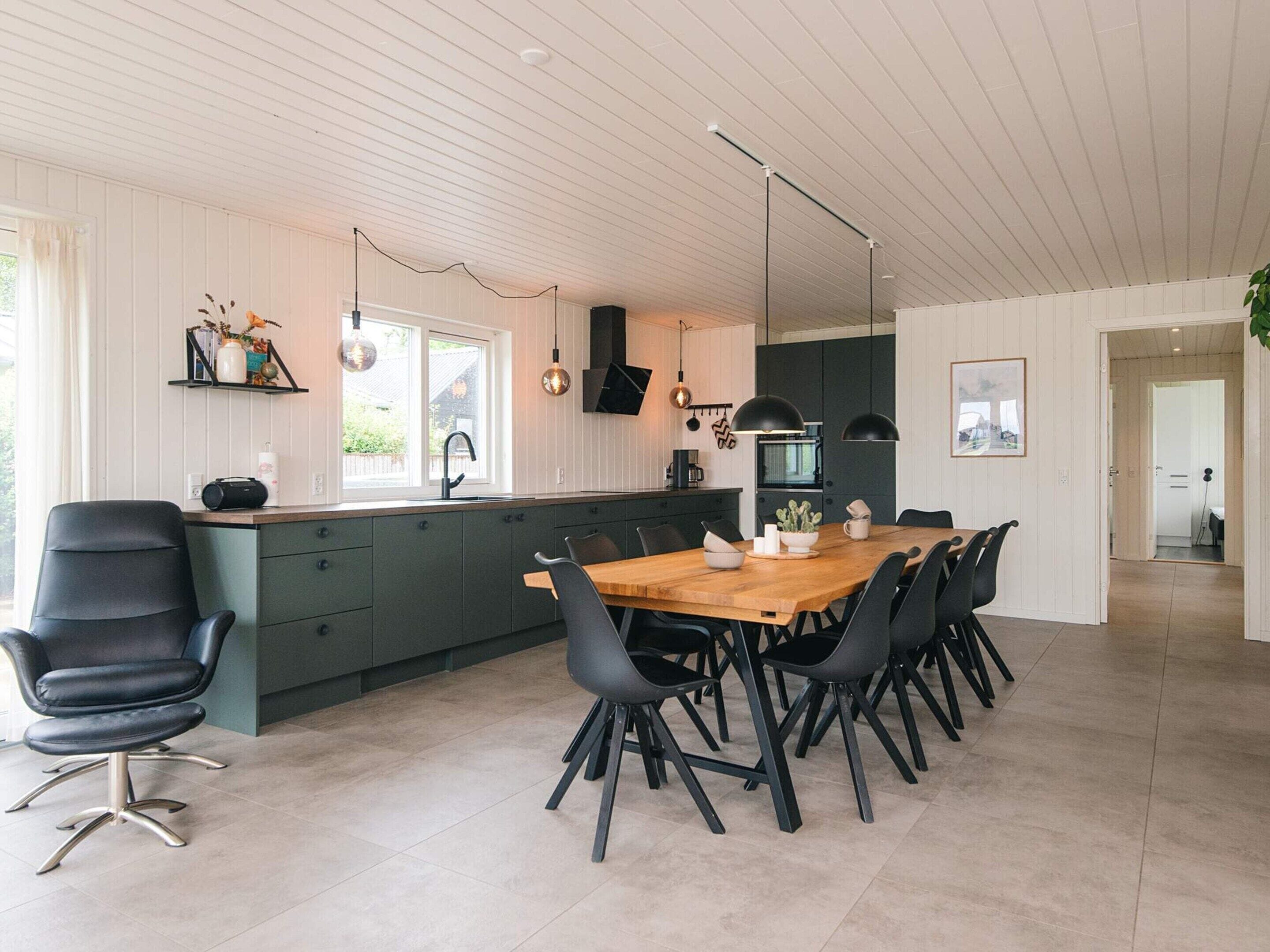Une cuisine moderne avec un îlot central, des chaises noires, une table à manger en bois, des luminaires suspendus et une vue sur l’extérieur.