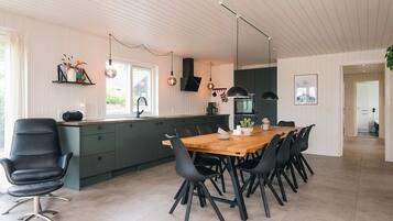 A modern kitchen with a central island, black chairs, a wooden dining table, pendant lights, and a view of the outdoors.
